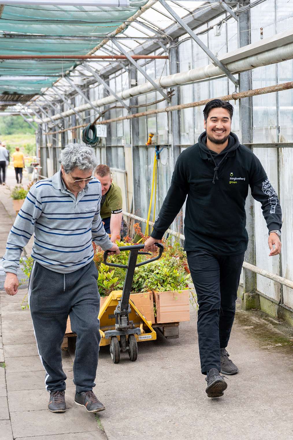 Jonge man trekt samen met oudere man een kar met planten in tuinkas, Ergotherapie HvA