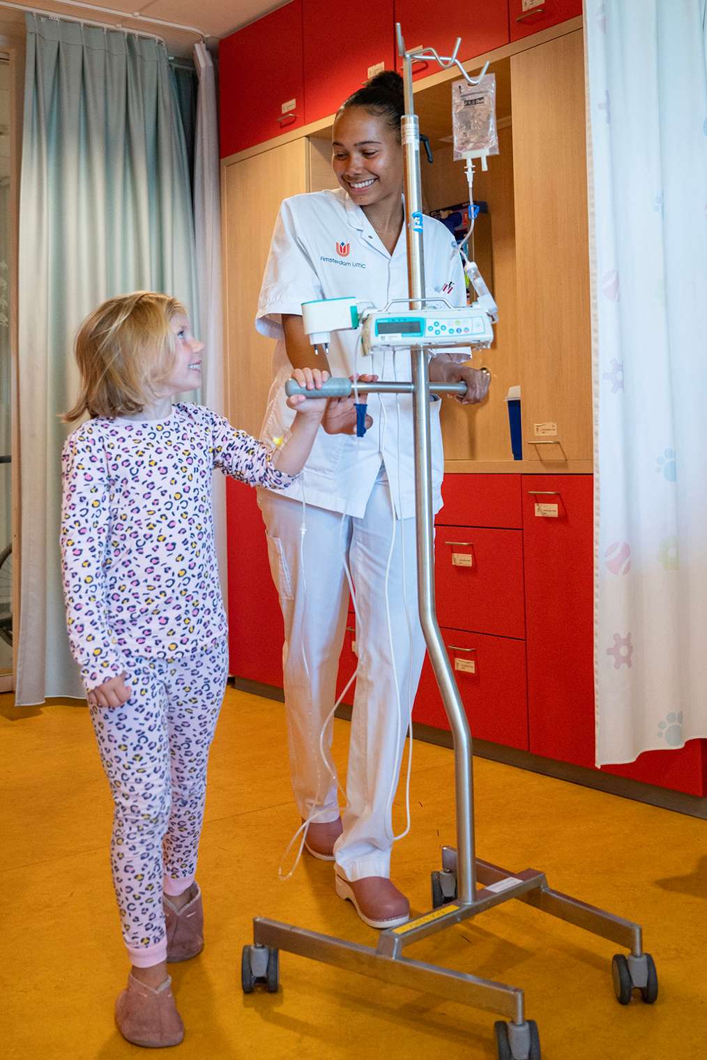 Verpleegkundige loopt met jonge patiënt en infuusstandaard door gang ziekenhuis